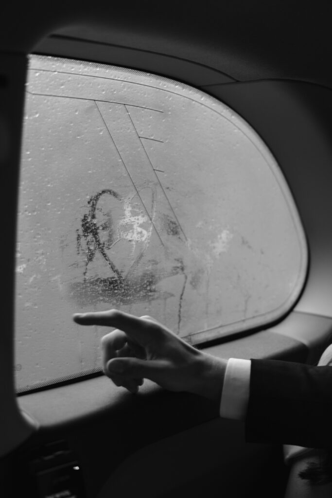 A person in a suit traces a drawing on a foggy car window with their finger on a rainy day. The window has water droplets and a sketch of a face is visible on the glass.