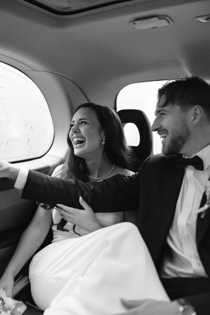 A joyful bride and groom sit together in the backseat of a car, smiling and laughing. The bride reaches toward the window while the groom sits beside her, both dressed in formal wedding attire.