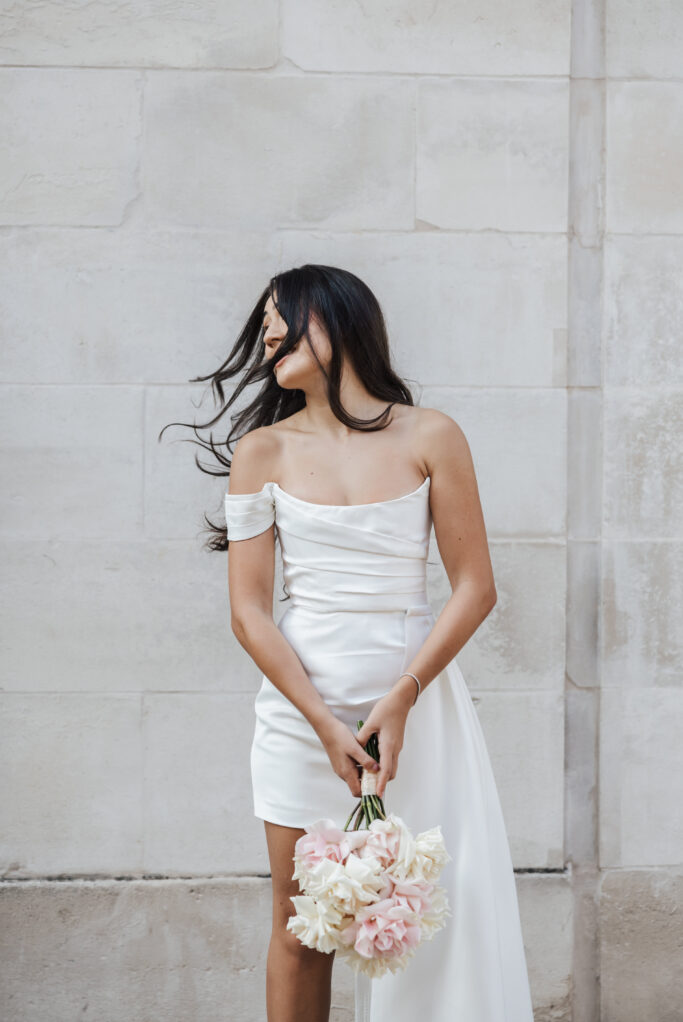 A woman in an off-shoulder white dress holds a bouquet of flowers and stands against a light stone wall. Her long dark hair is blowing across her face as she looks to the side.