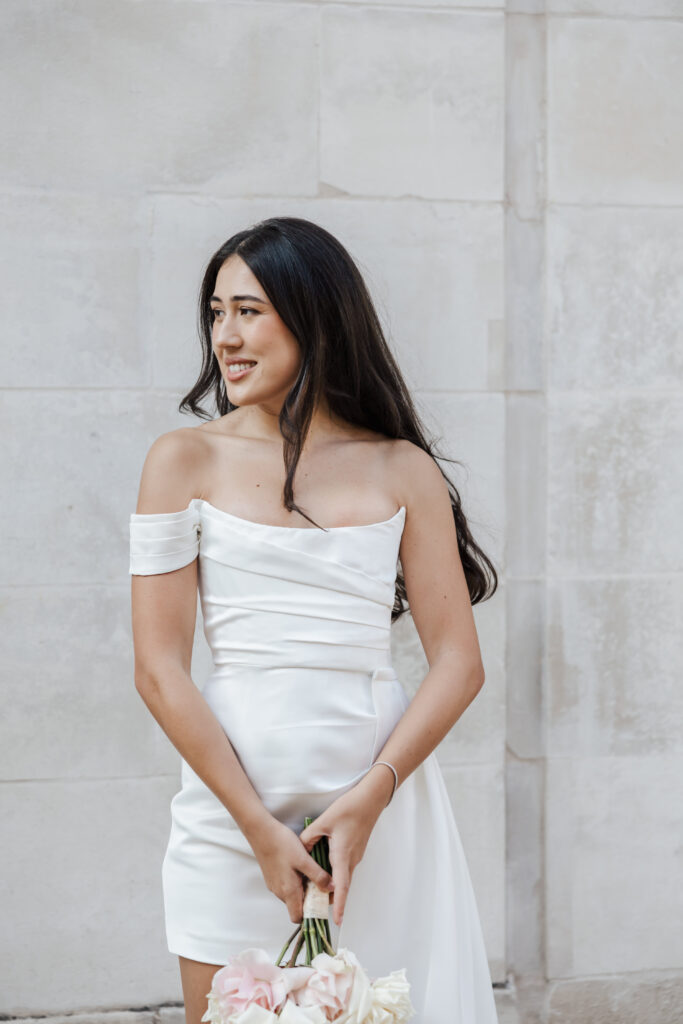 A woman in a white, off-the-shoulder dress stands against a light stone wall, holding a bouquet of flowers and smiling while looking to her left.