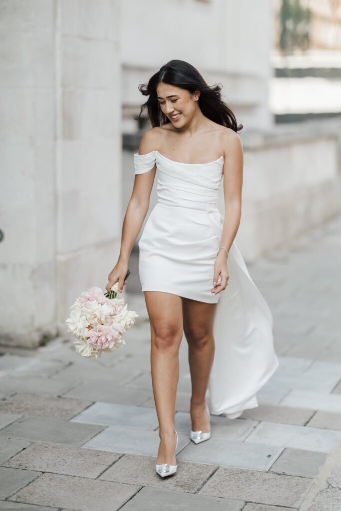 A woman in an off-shoulder white dress holds a bouquet of pale pink and white flowers, smiling as she walks along a city sidewalk.