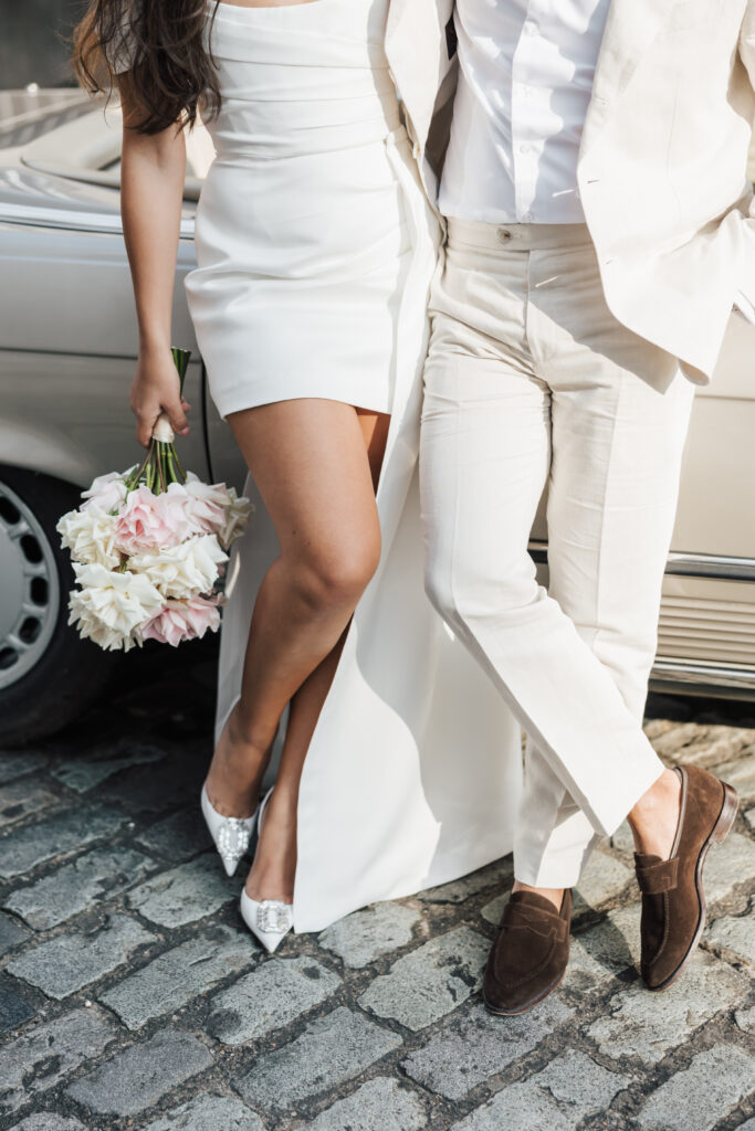 A couple dressed in white wedding attire stands next to a vintage car on a cobblestone street. The woman holds a bouquet of flowers, and both have their legs crossed, with their faces out of the frame.
