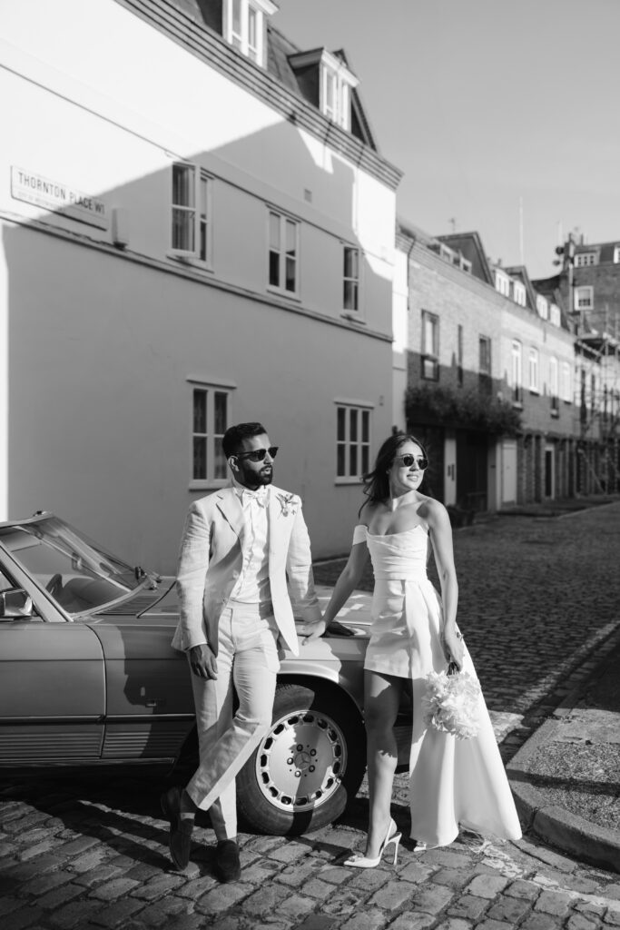A stylish couple in wedding attire stand beside a vintage car on a cobblestone street. The man wears a light suit and sunglasses; the woman wears a short dress with a train, holding a bouquet, both smiling in the sunlight.