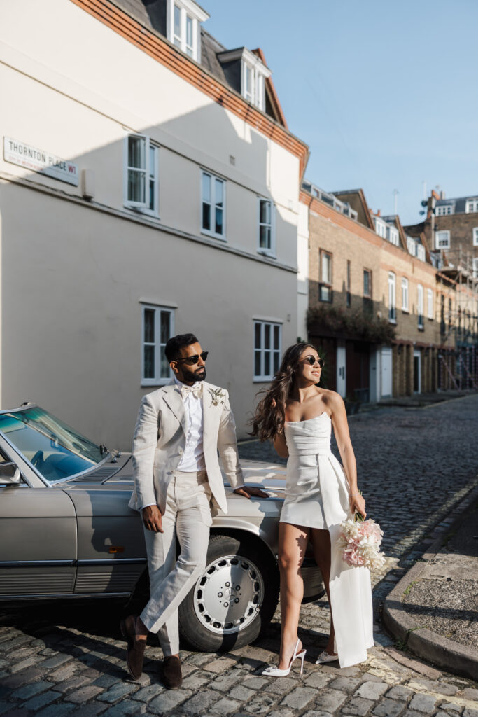 A stylish couple dressed in white wedding attire leans against a classic silver car on a cobblestone street, holding a bouquet of flowers and wearing sunglasses, with buildings in the background.