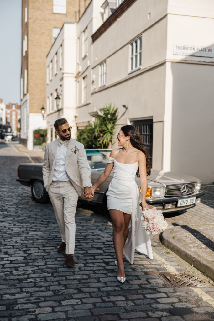 A couple in wedding attire hold hands and walk along a cobblestone street. The groom wears a light suit and sunglasses, while the bride wears a short white dress and holds a bouquet. A vintage car is parked behind them.