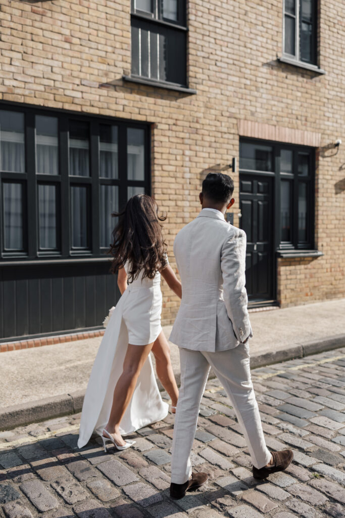 A couple dressed in light-colored formal wear walks hand in hand down a cobblestone street beside a brick building with black-trimmed windows. The woman wears a white dress and heels; the man wears a light suit and brown shoes.