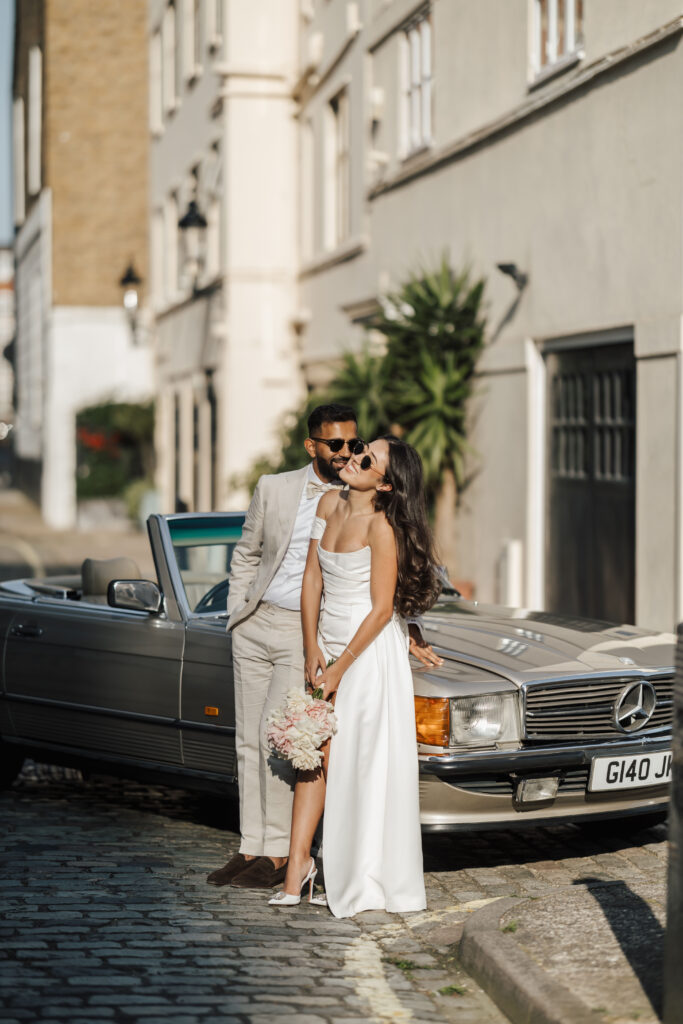 A bride in a white dress and a groom in a light suit stand by a silver convertible Mercedes on a sunlit street. The bride holds a bouquet and both appear happy, wearing sunglasses and smiling.