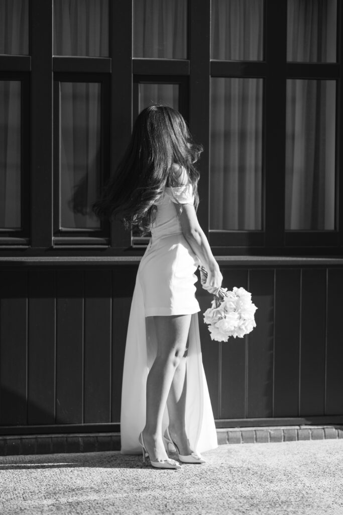 A woman in a short, off-shoulder white dress and high heels stands outdoors, holding a bouquet of flowers, with her long hair swaying as she turns away from the camera.