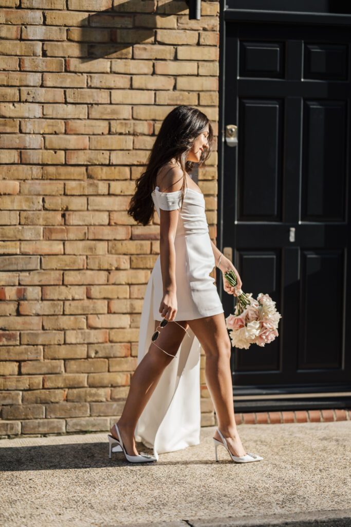 A woman in a white off-shoulder dress and high heels walks outside on a sunny day, holding a bouquet of pale pink flowers, with a brick wall and black door in the background.