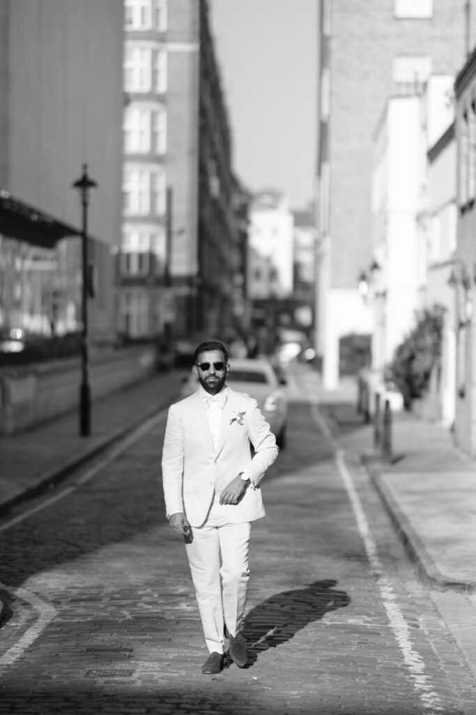 A man in a light-colored suit and sunglasses walks confidently down a cobblestone city street, with tall buildings on either side and sunlight casting shadows behind him.