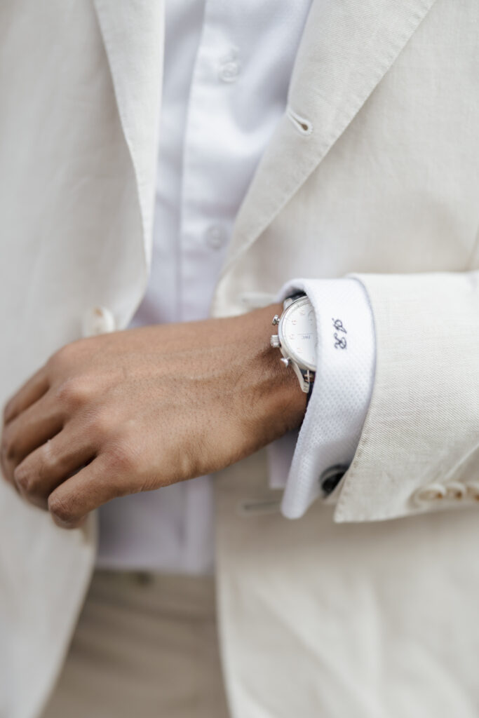 A person wearing a white suit and white shirt displays a silver wristwatch on their left wrist. The cuff is monogrammed with the initials GP. The image focuses on the hand and lower arm.