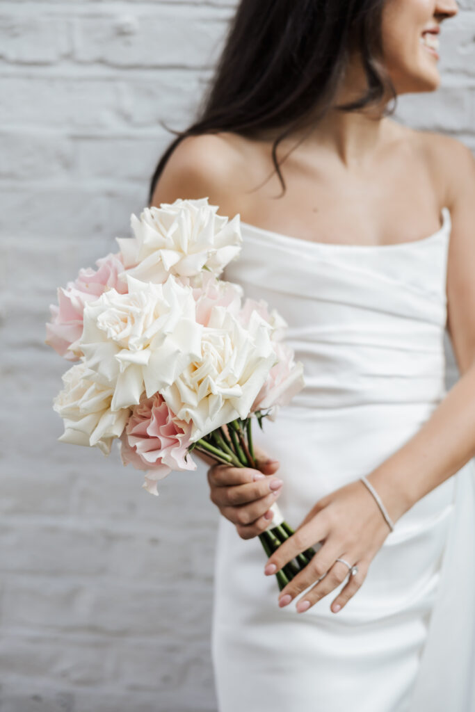A woman in a strapless white dress holds a bouquet of white and pale pink roses, smiling and standing against a light brick wall. Her hair is loose, and she is wearing a ring and a simple bracelet.