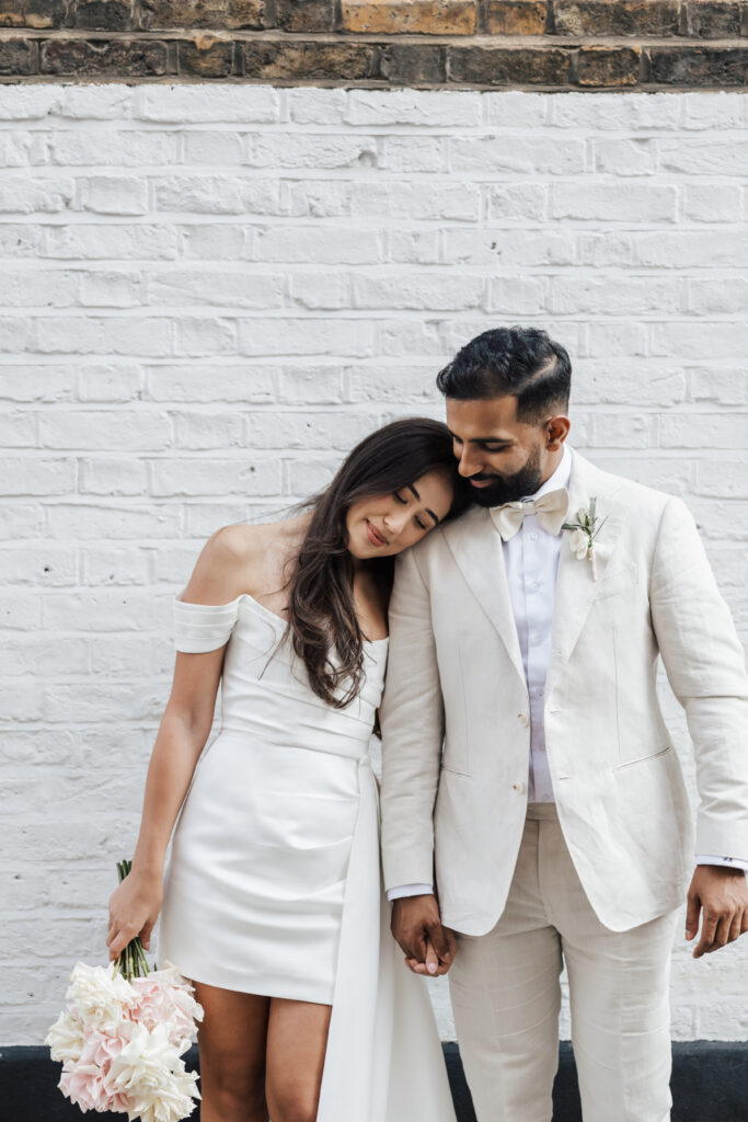 A bride in an off-shoulder white dress leans her head on a groom’s shoulder. The groom wears a light suit with a bow tie. They hold hands and the bride carries a bouquet, standing against a white brick wall.