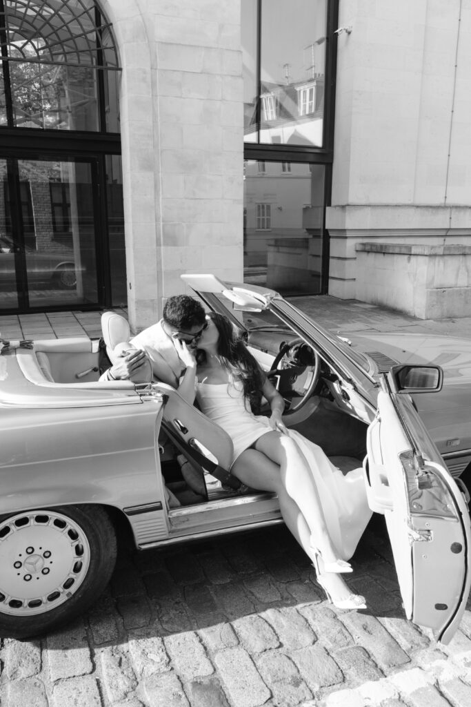 A couple dressed in wedding attire share a kiss while sitting in a convertible car with the door open, parked on a cobblestone street in front of a building. The scene is in black and white.
