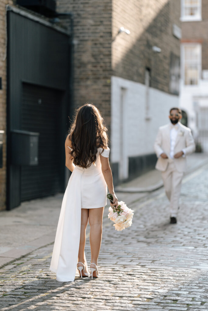 A woman in a short white dress holding a bouquet stands on a cobblestone street, facing a man in a light suit and sunglasses walking toward her. The scene is outdoors with brick buildings.