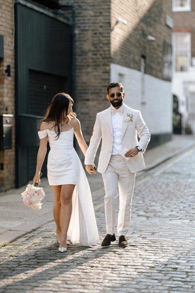 A couple dressed in elegant white wedding attire walk hand-in-hand down a cobblestone street. The woman holds a bouquet of flowers and wears an off-shoulder dress, while the man wears a light suit and sunglasses.