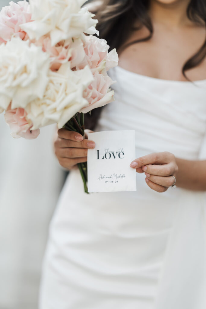 A bride in a white dress holds a bouquet of pale pink roses in one hand and a small card with the word Love and names written on it in the other hand. Her face is not visible.