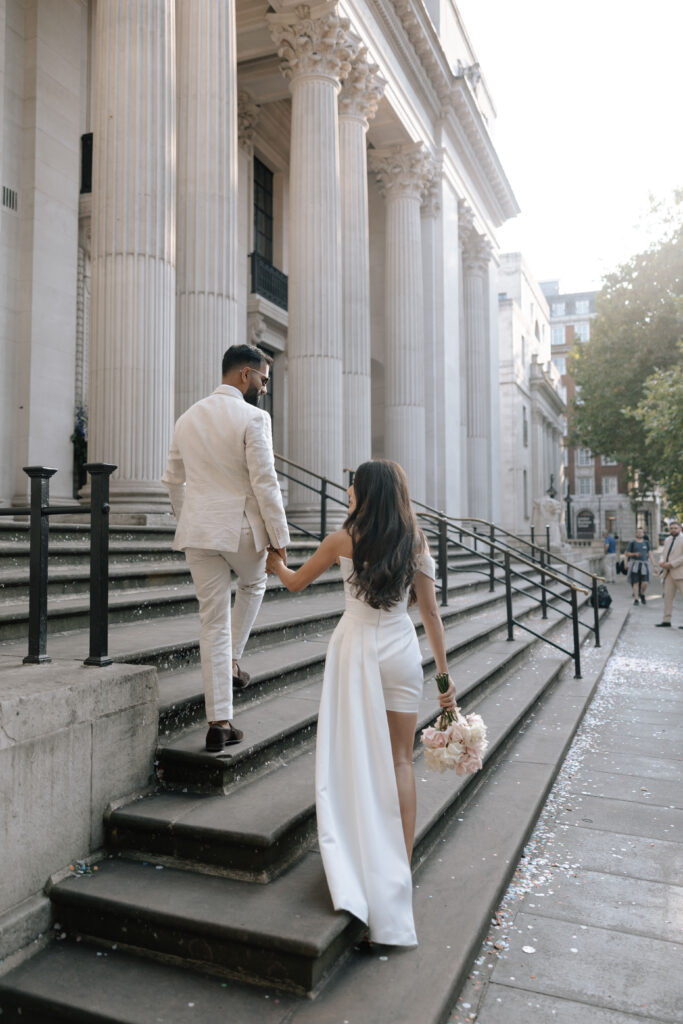 A couple in white formal attire walk up stone steps outside a grand building with tall columns; the woman holds a bouquet of flowers and wears a long dress.