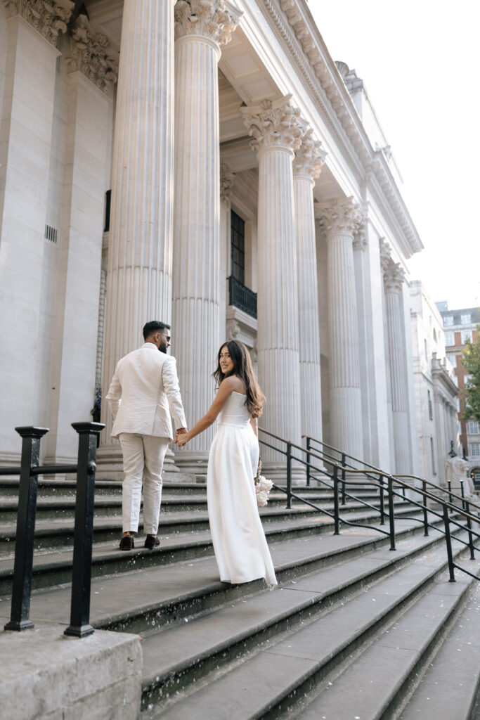 A couple dressed in elegant white attire holds hands while walking up the steps of a grand building with tall columns and ornate architecture. The woman looks back while ascending the stairs.