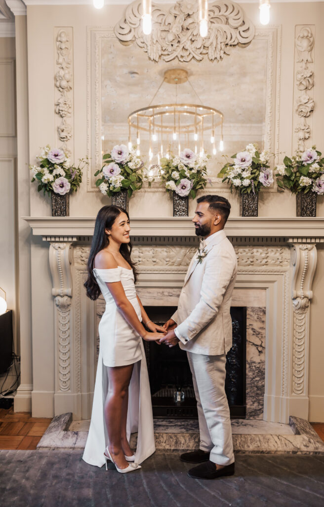 A couple dressed in white stands holding hands in front of an ornate fireplace decorated with flowers, facing each other and smiling in a warmly lit room.