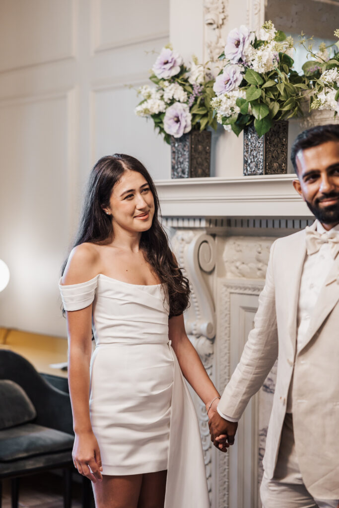 A woman in a white off-shoulder dress holds hands with a man in a cream suit, standing by an ornate fireplace decorated with vases of purple and white flowers. She is smiling softly while looking at him.
