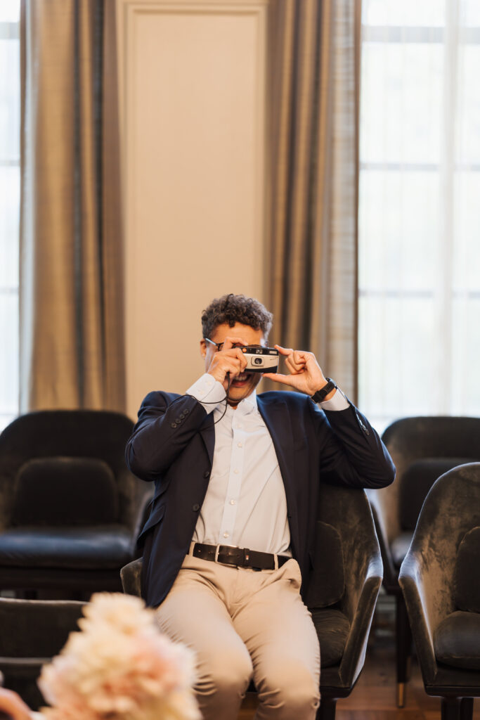 A man in a navy blazer and light pants sits on a chair indoors, holding a camera up to his face as he takes a photo. The background features empty chairs and large, sunlit windows with curtains.