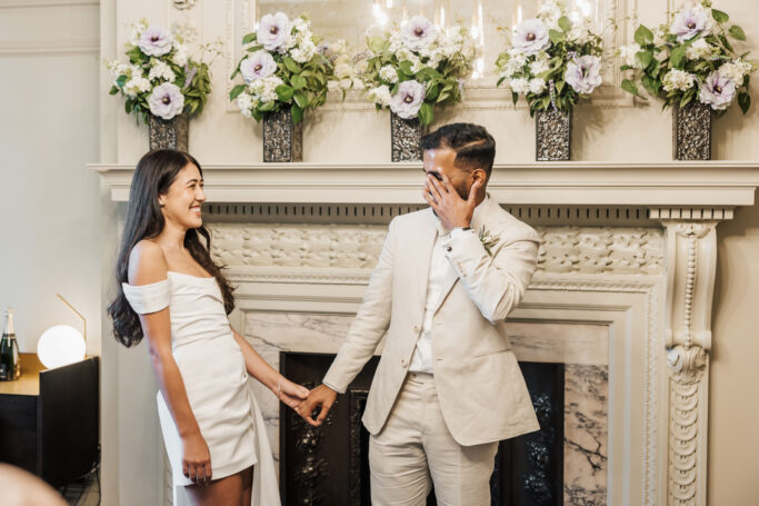 A couple in white outfits stands in front of a decorated fireplace, holding hands. The woman smiles, while the man wipes a tear from his eye. Flower arrangements are displayed above the mantle.