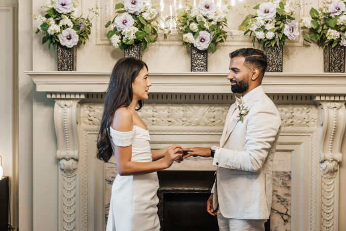 A bride and groom exchange rings in front of a decorated fireplace adorned with white and purple flowers during an indoor wedding ceremony.