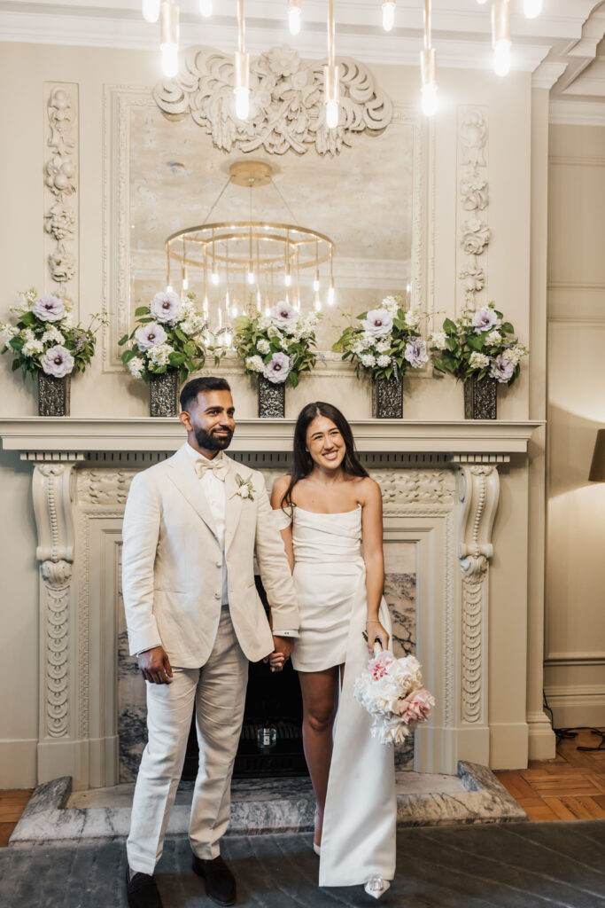 A couple dressed in white stands smiling and holding hands in front of a decorated fireplace with flowers and a mirror, under modern hanging lights in an elegant room. The woman holds a bouquet.