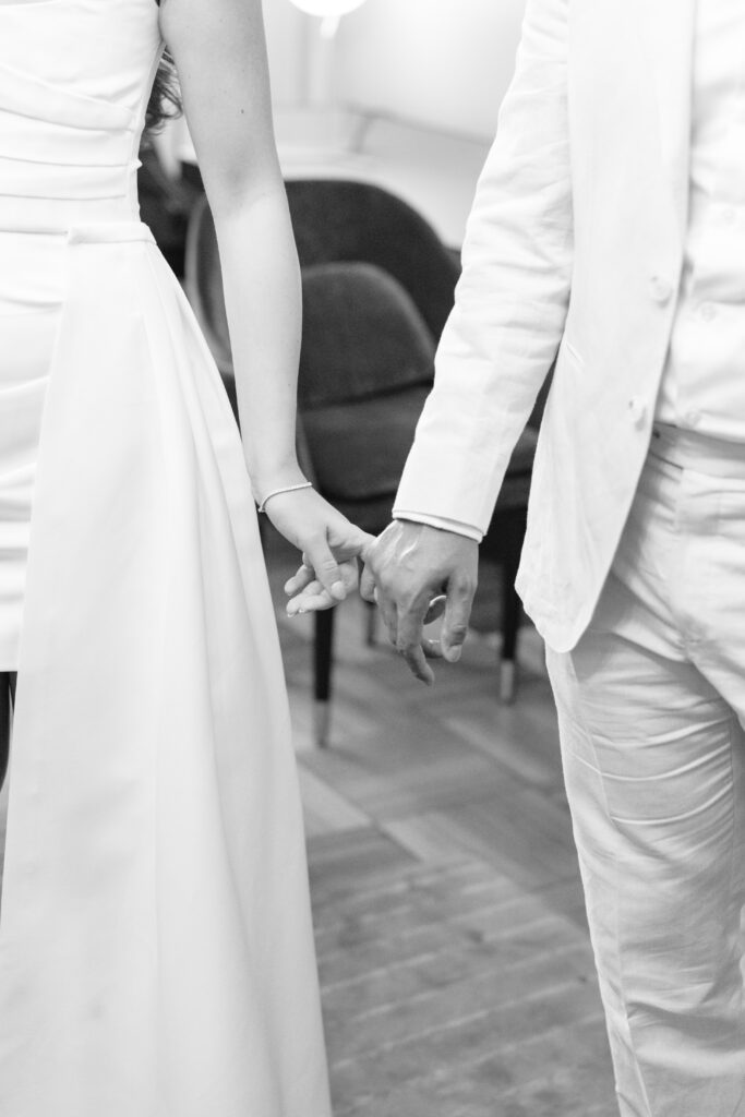 A black and white photo showing a couple in formal attire holding hands; only their torsos and arms are visible, with both wearing light-colored clothing.