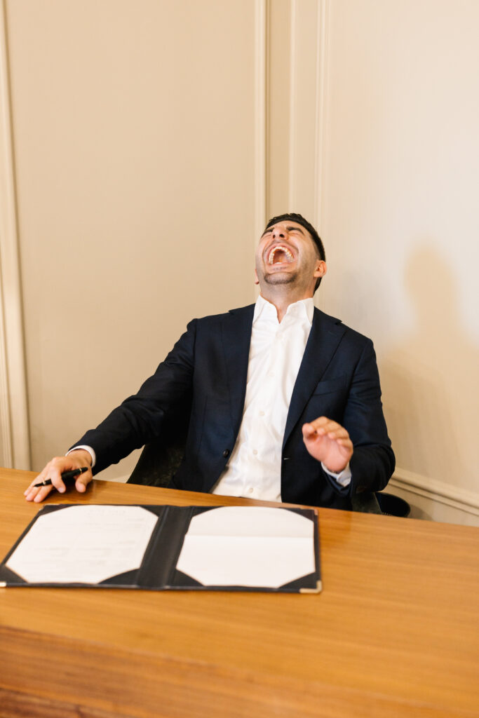 A man in a suit sits at a desk with an open folder of papers, leaning back and laughing with his head tilted up and eyes closed, appearing joyful and carefree.