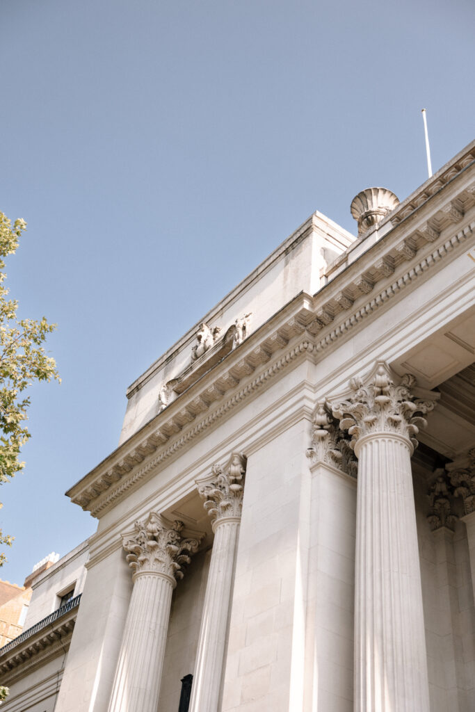 White neoclassical building with tall Corinthian columns, decorative carvings, and sculptures on the roof, set against a clear blue sky with part of a leafy tree visible on the left.