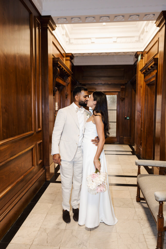 A couple dressed in elegant white attire shares a loving moment in a hallway with wooden walls. The woman holds a bouquet of flowers, and they look into each others eyes, smiling gently.