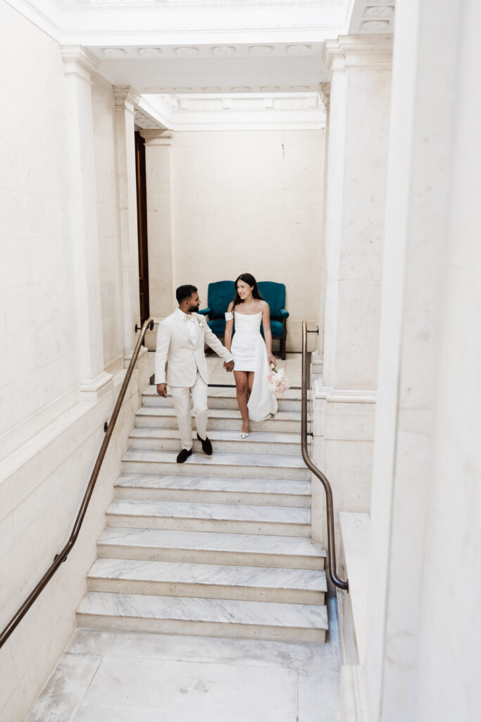 A couple dressed in elegant white attire walks hand in hand down marble stairs in a bright, modern building with white walls and a teal chair in the background. The woman holds a bouquet of flowers.