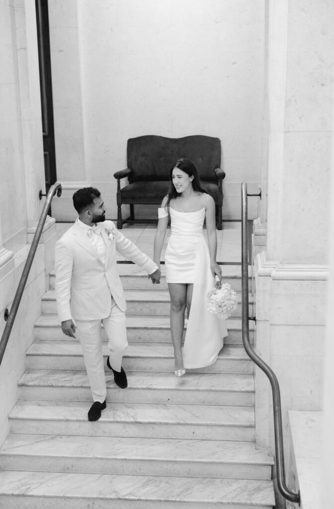 A couple in white formal attire walks down marble stairs, holding hands. The woman carries a bouquet and wears a one-shoulder dress; the man wears a suit with a bow tie. The setting appears elegant and classic.