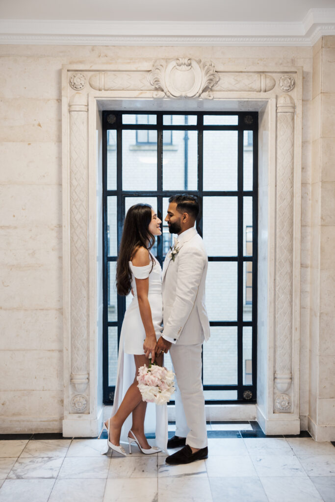 A couple dressed in white stands face to face, holding hands and a bouquet of flowers, in front of a large window with a black grid frame, set in a marble-walled room.