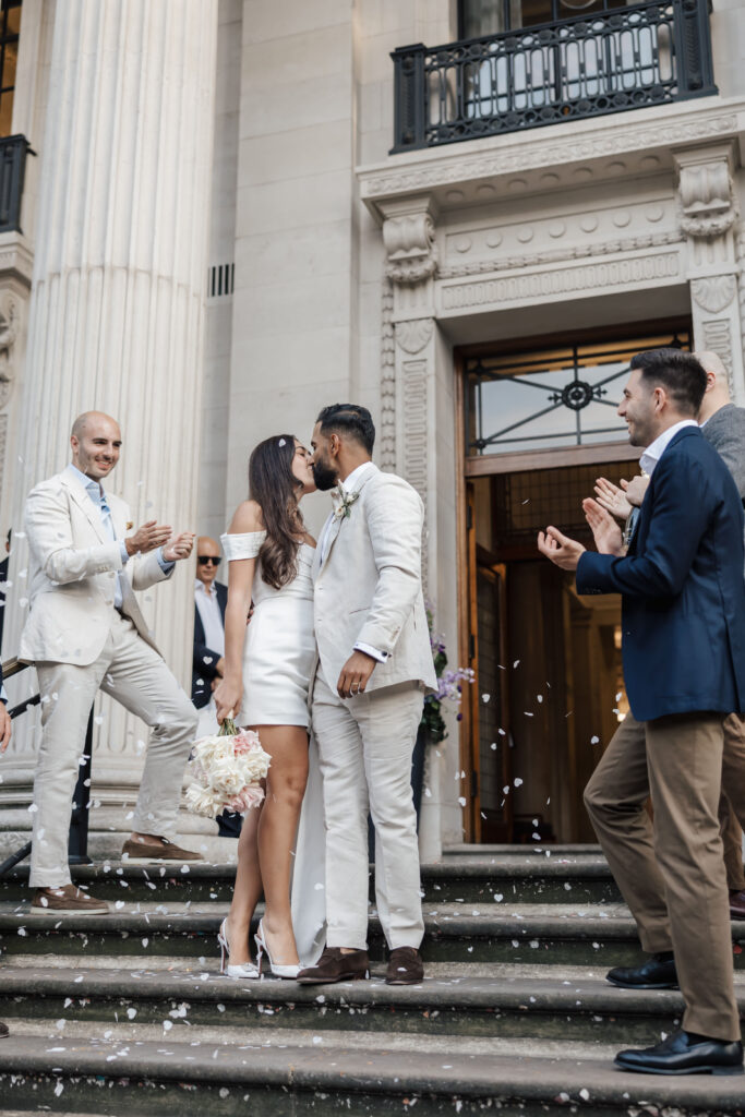 A newlywed couple kisses on the steps of a grand building while friends in suits clap and toss confetti. The bride wears a short white dress and heels, and the groom is in a light-colored suit.