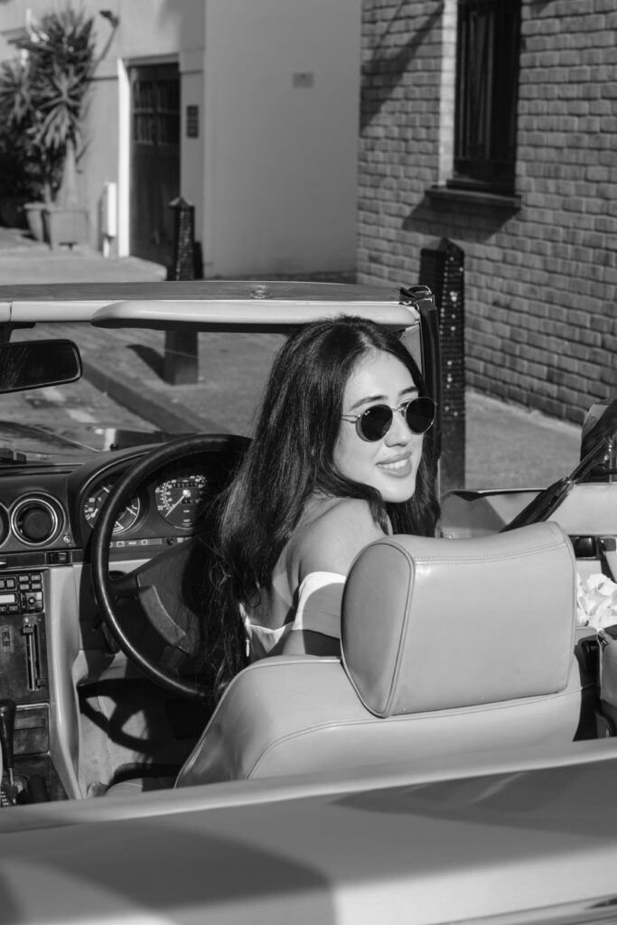 A woman with long dark hair and sunglasses smiles while sitting in the driver’s seat of a convertible car, looking back over her shoulder. The photo is black and white and taken on a sunny day in an urban setting.
