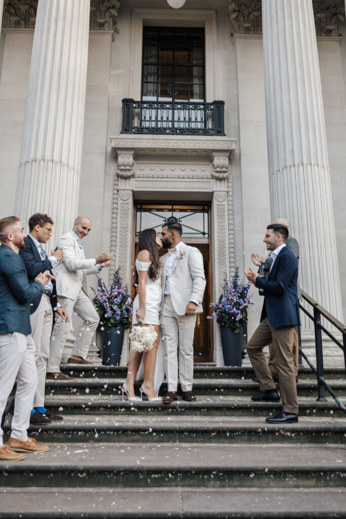 A bride and groom stand on steps outside a grand building, surrounded by well-dressed guests clapping and celebrating. The bride holds a bouquet and kisses the groom as white confetti falls around them.