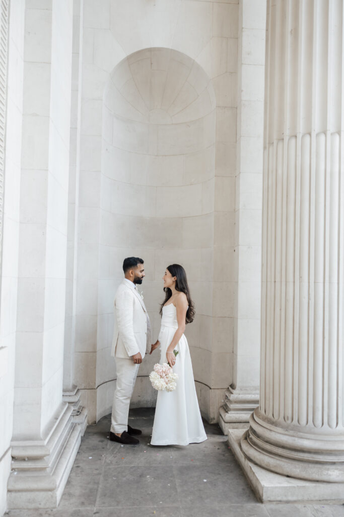 A bride and groom stand facing each other in a white, columned architectural setting. The bride holds a bouquet of flowers and wears a long white dress; the groom wears a light-colored suit.