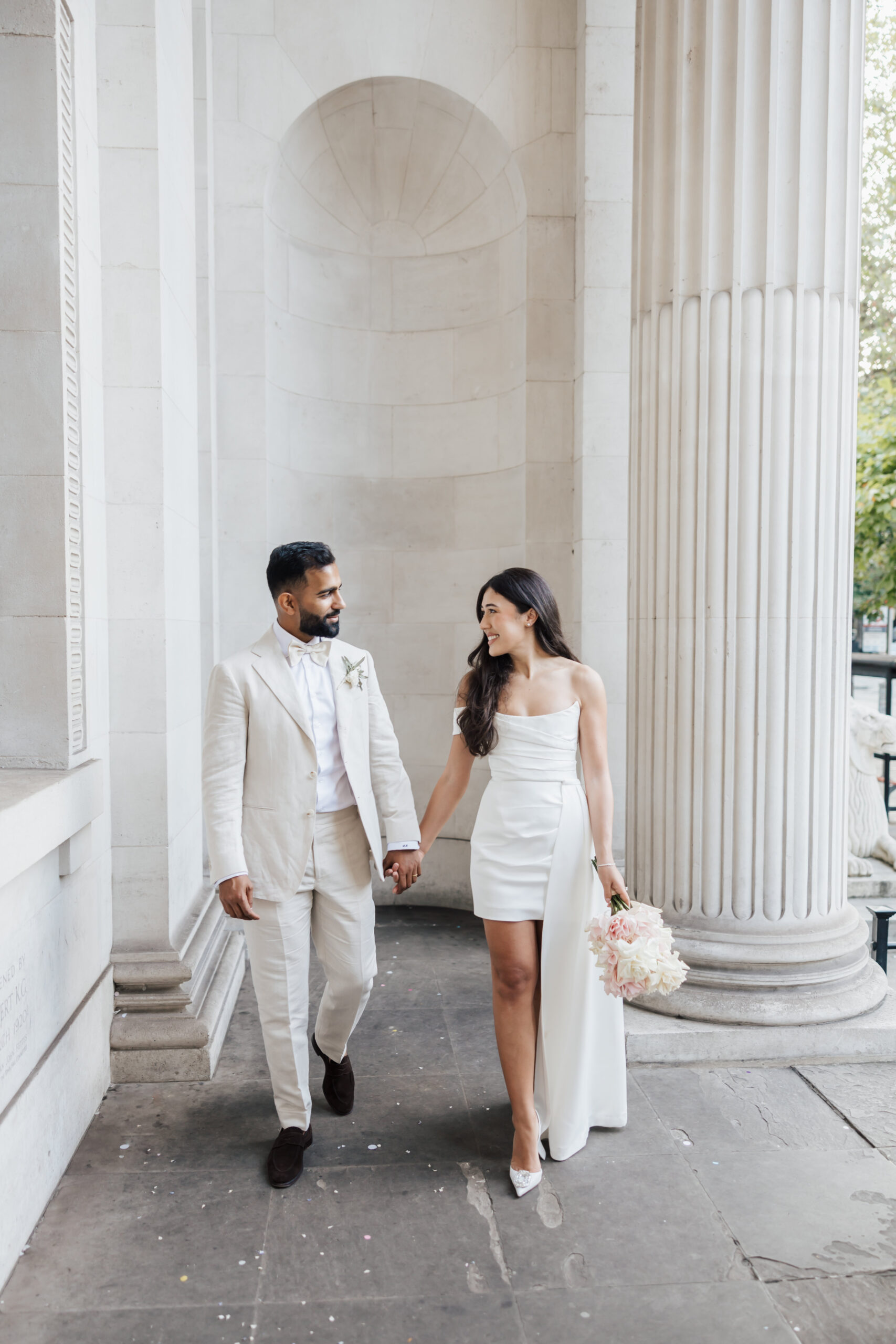 A couple in wedding attire walks hand in hand outside a neoclassical building with tall columns. The groom wears a light beige suit; the bride wears a short white dress and holds a bouquet of flowers. Both are smiling at each other.
