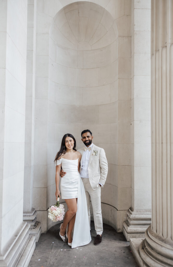 A couple dressed in wedding attire stands closely together in an elegant, stone archway. The woman holds a bouquet and wears a short white dress; the man wears a light-colored suit with a boutonniere. Both are smiling.