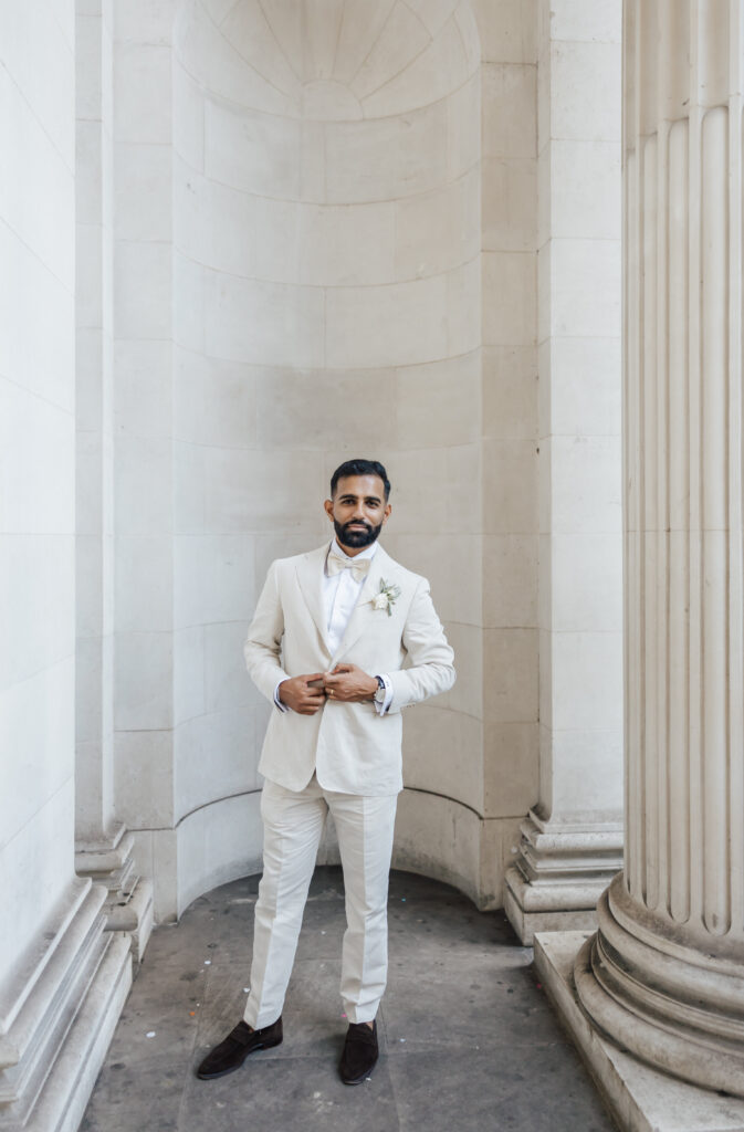 A man with a beard, dressed in a white suit and white shirt, stands confidently between large, white stone columns and walls. He adjusts his jacket and has a boutonniere on his lapel.