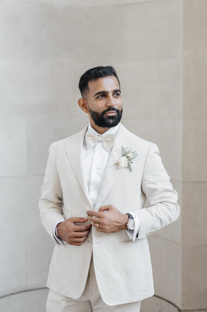 A man with a beard, wearing a cream-colored suit, white shirt, and bow tie, stands against a light stone wall. He has a white boutonnière on his lapel and is adjusting his jacket, looking to the side and smiling slightly.