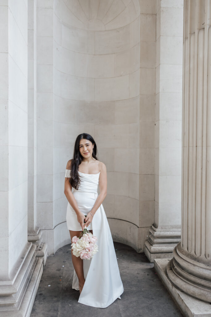 A woman in a white, off-the-shoulder dress holds a bouquet of flowers and stands between large stone columns with a curved wall behind her. She has long dark hair and is smiling gently.