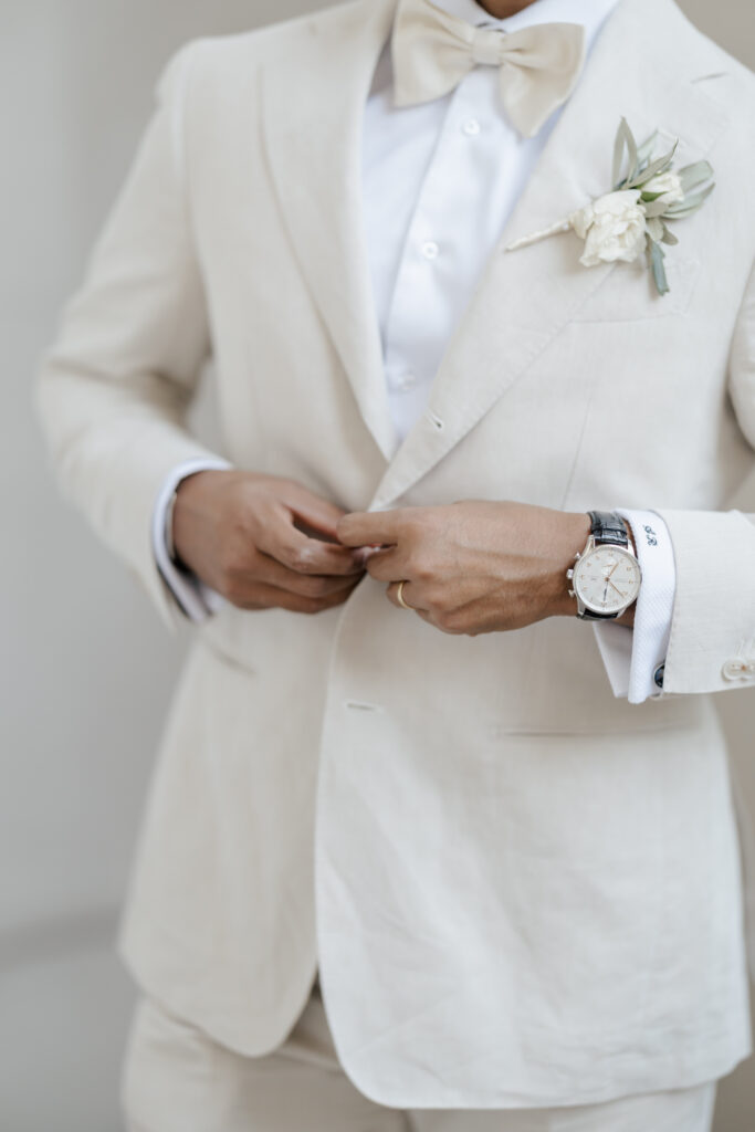 A person in a cream-colored suit with a white shirt and bow tie buttons their jacket. They wear a watch and a wedding ring, and have a white boutonniere pinned to the lapel. The face is not visible.
