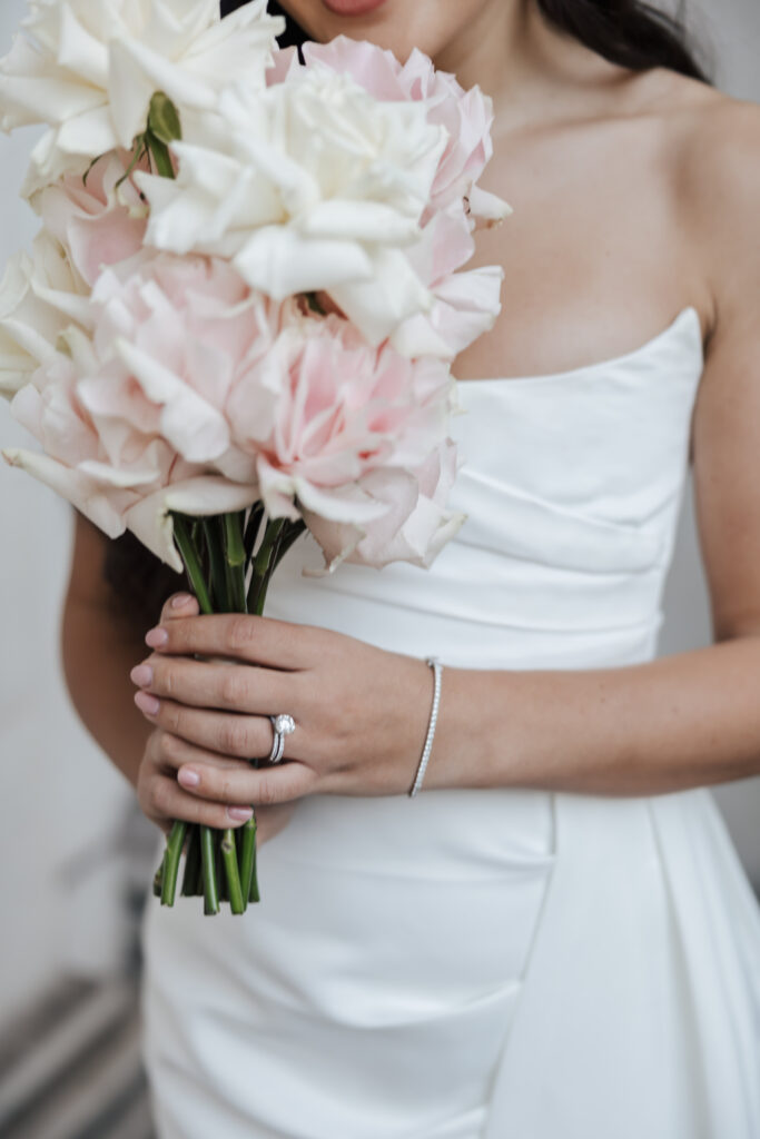 A bride in a strapless white dress holds a bouquet of white and pale pink roses. She wears a silver ring and bracelet, and her long, dark hair cascades over her shoulder.