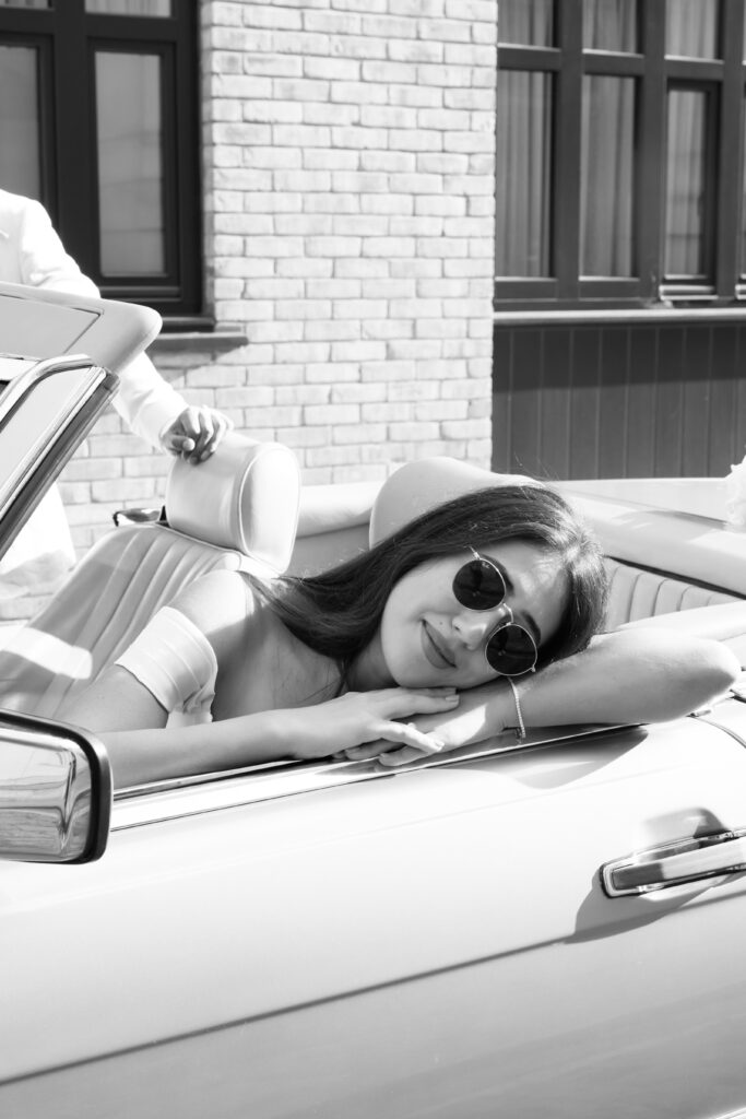 A woman wearing sunglasses leans her head and arms on the door of a convertible car, smiling softly. The photo is in black and white, with a brick building in the background.