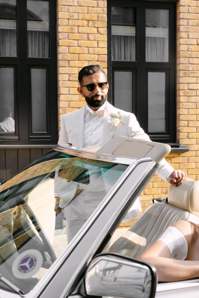 A man in a white suit and sunglasses stands beside a white convertible car, looking away. The background features yellow brick walls and black-framed windows. Another person is seated in the car, mostly out of frame.