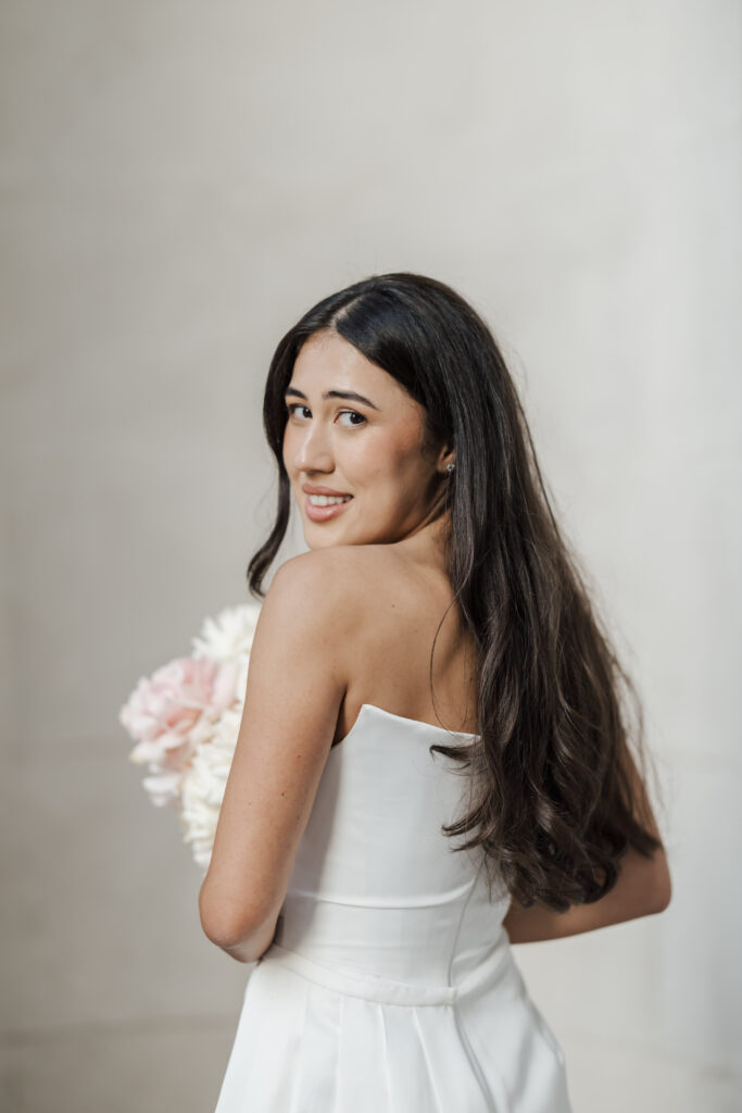 A woman in a white dress holding a bouquet of light pink and white flowers looks over her shoulder and smiles at the camera, standing against a light, neutral background.